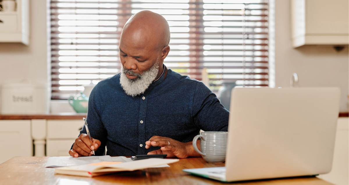 A man writing in a notebook in front of a laptop computer